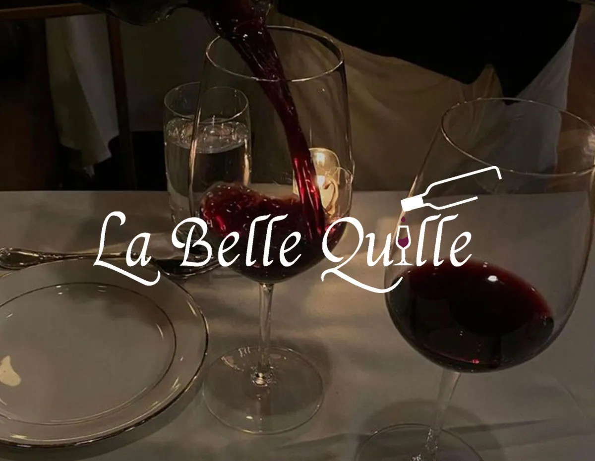 Logo La Belle Quille sur une photo d'un serveur versant du vin rouge dans un verre sur une table dressée.