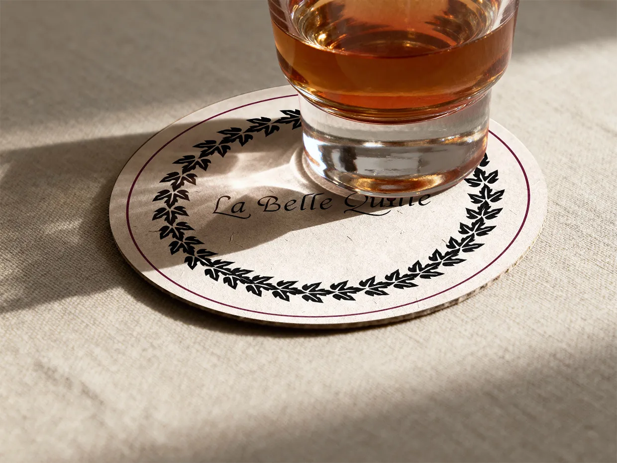 Mockup d'un sous-verre en carton avec le logo La Belle Quille et une couronne de feuilles, sous un verre de spiritueux ambré.