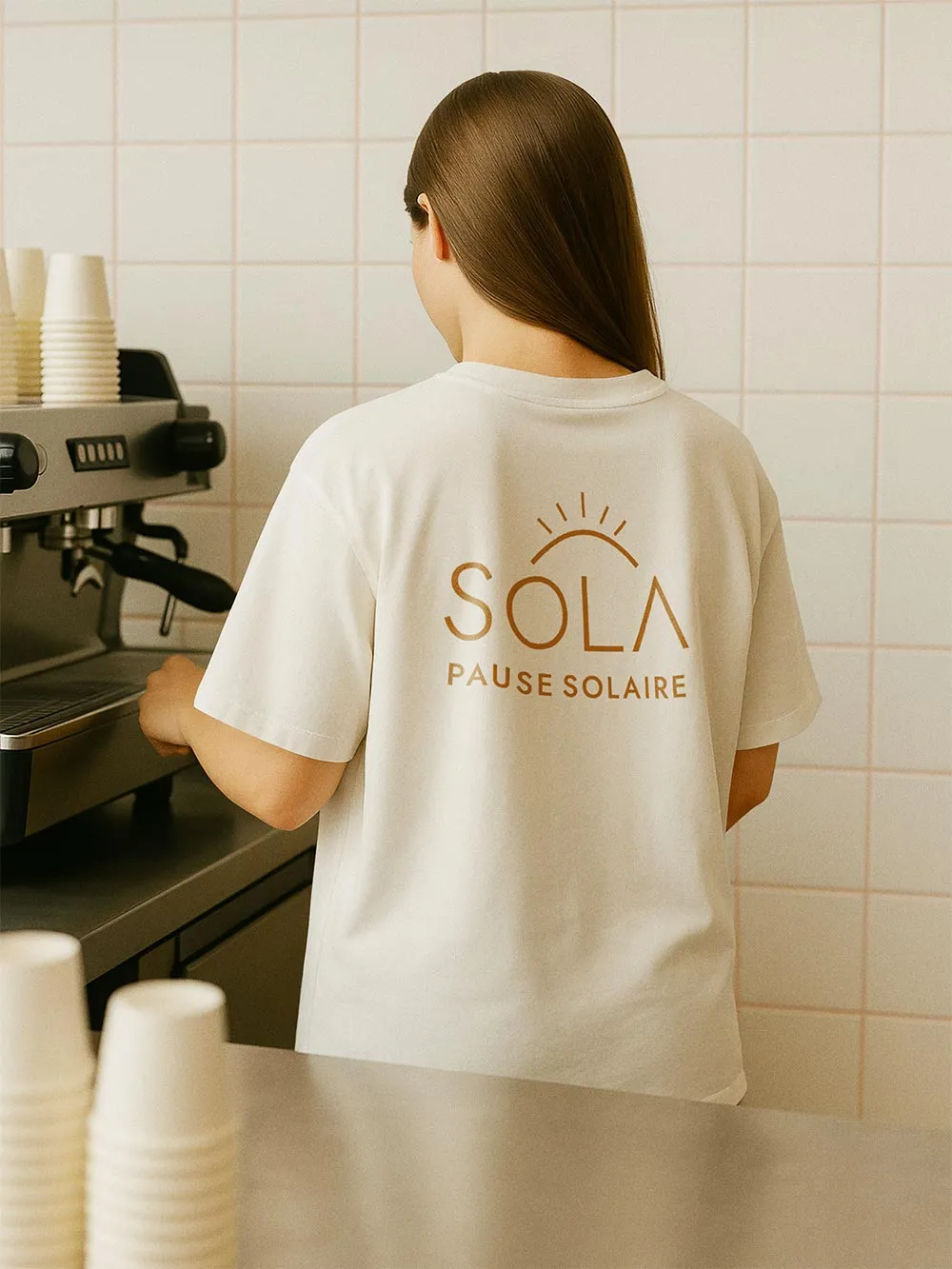 Dos d'une femme portant le t-shirt blanc SOLA avec le logo ocre et la baseline Pause Solaire dans un environnement de café.