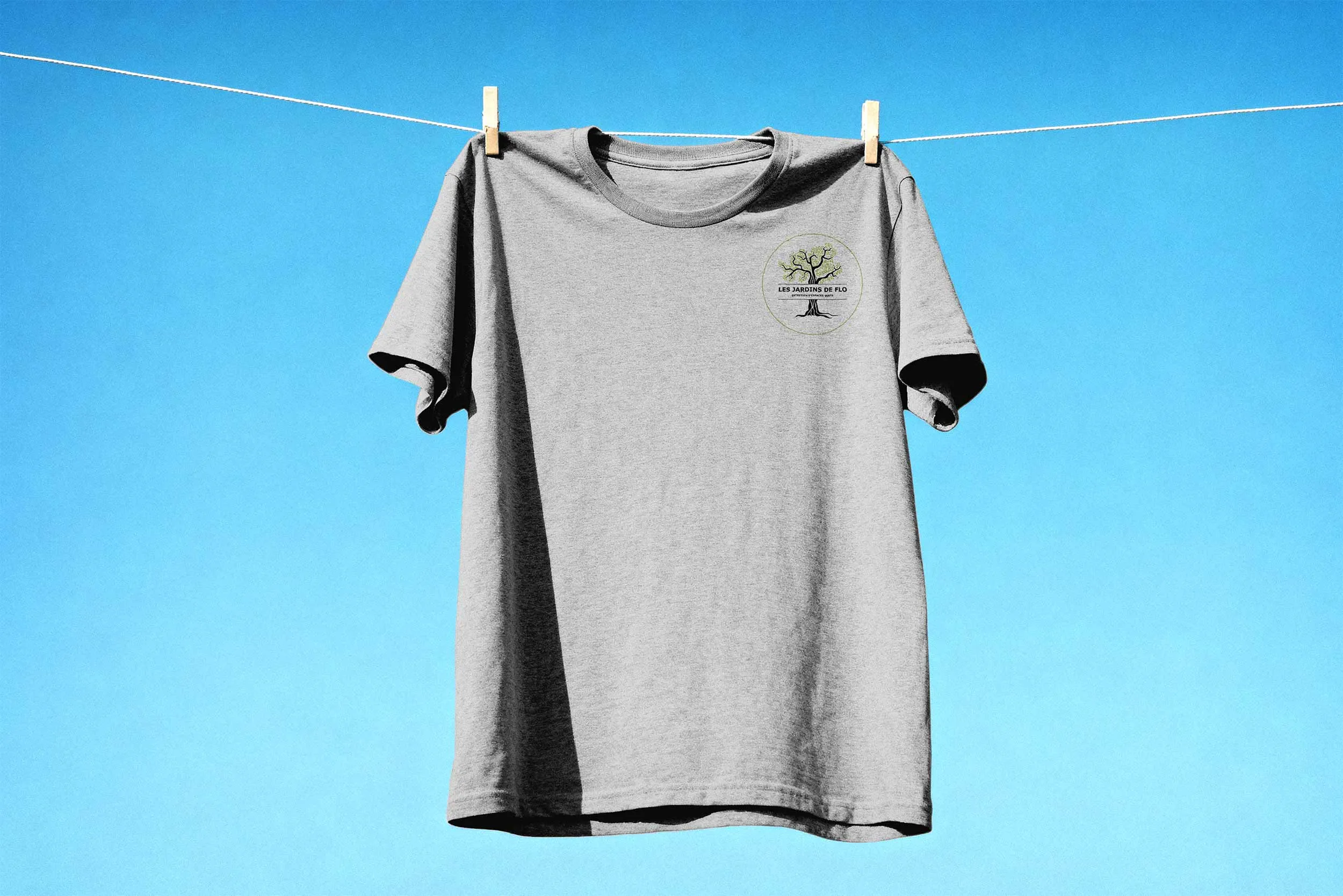 T-shirt gris avec le logo des Jardins de Flo imprimé côté cœur, suspendu sur un fil à linge sous un ciel bleu.
