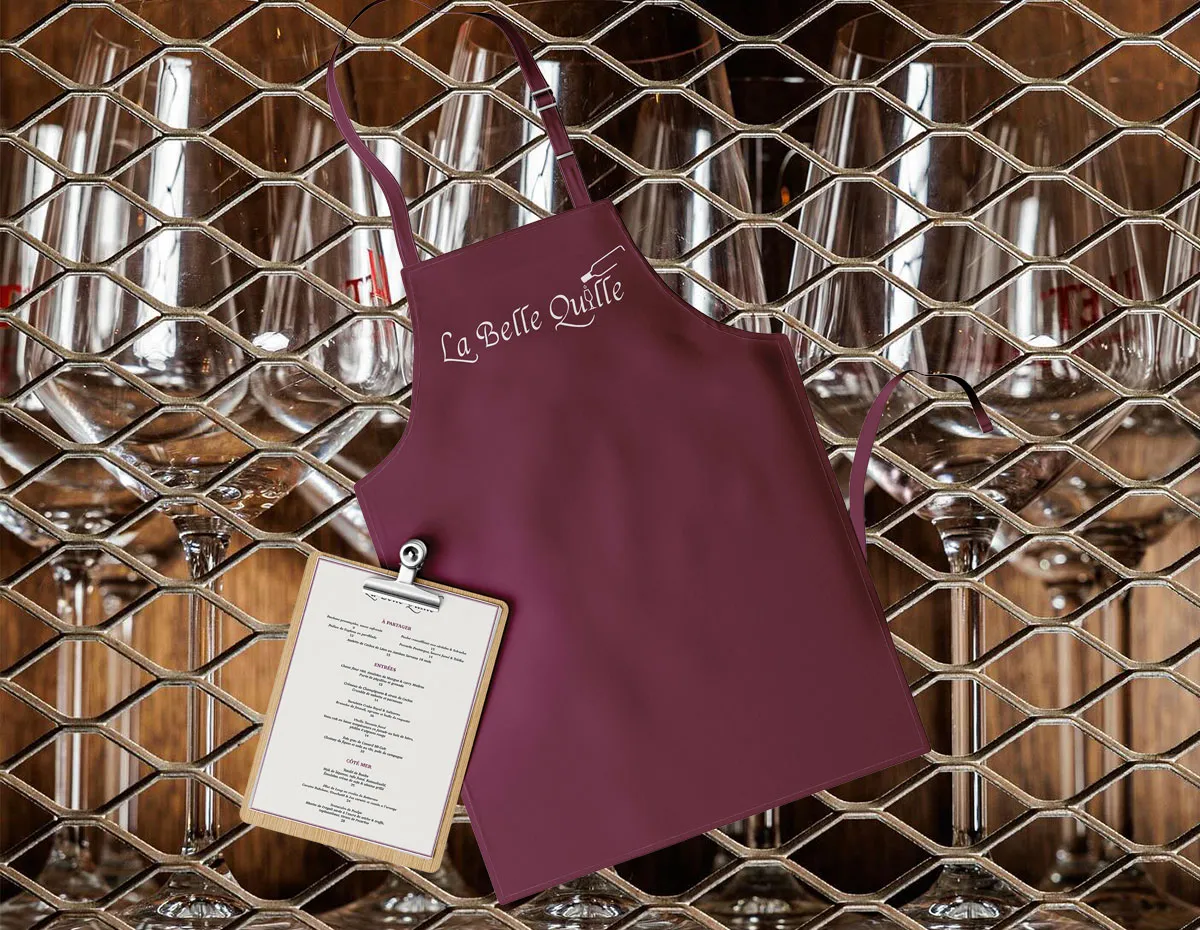 Tablier bordeaux avec logo blanc La Belle Quille et menu sur un porte-bloc devant un rack de verres à vin.